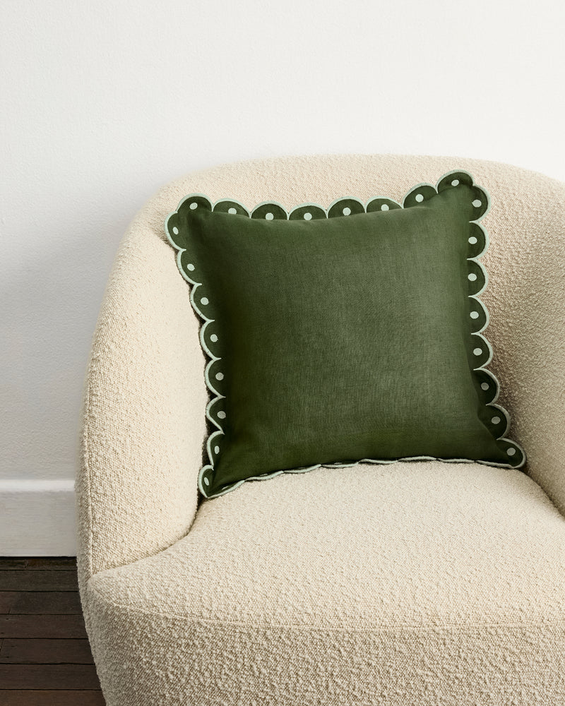 A plush, cream armchair holds the Bed Threads Olive & Sage 100% French Flax Linen Arch Cushion Cover—dark green with scalloped edges and white embroidery—set against a white wall and dark wooden floor.