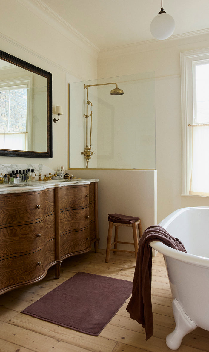 A bright bathroom with a freestanding white bathtub, a brown towel draped over its edge, a wooden vanity with a marble top and mirror, a brown mat, and a glass-enclosed shower with gold fixtures.