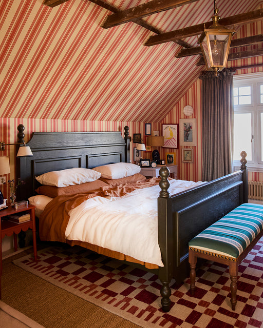A cozy bedroom features a black wooden bed, brown and white bedding with Bed Threads’ Rust 100% French Flax Linen Pillowcases, a striped bench, red striped wallpaper, exposed ceiling beams, and a large window filling the space with natural light.