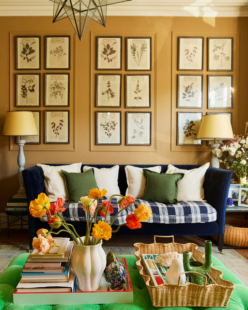 A cozy living room with a blue velvet sofa, Bed Threads Olive 100% French Flax Linen Cushion Covers in white and green, botanical prints on tan walls, two lamps, a coffee table with books, flowers in a vase, and decorative trays with ceramics.