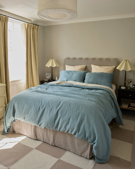 A cozy bedroom features a double bed with the Bed Threads Lagoon & Crème 100% French Flax Linen Bedding Bundle, beige pillows, and two black nightstands with lamps. Light curtains frame a window, complemented by soft, neutral decor.