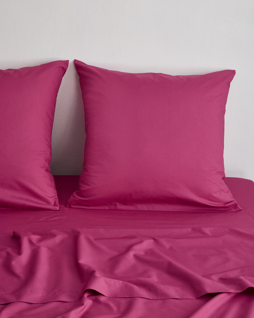 The Bed Threads Berry Organic Cotton European Pillowcases (Set of Two) and a matching magenta bed sheet are neatly displayed on a bed against a white wall, with soft folds visible in the sheet.
