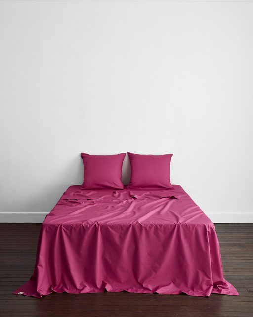 A neatly made bed features Berry Organic Cotton European Pillowcases (Set of Two) by Bed Threads in pink, set against a plain white wall on a dark wooden floor.