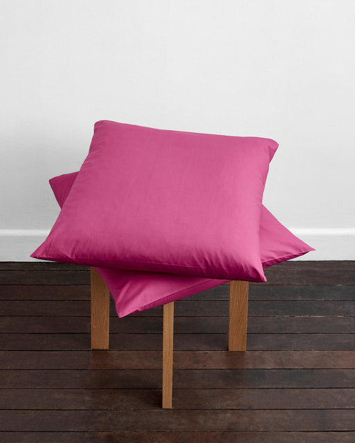 Two Berry Organic Cotton European Pillowcases by Bed Threads are stacked on a small wooden stool with a dark wooden floor and white wall in the background.