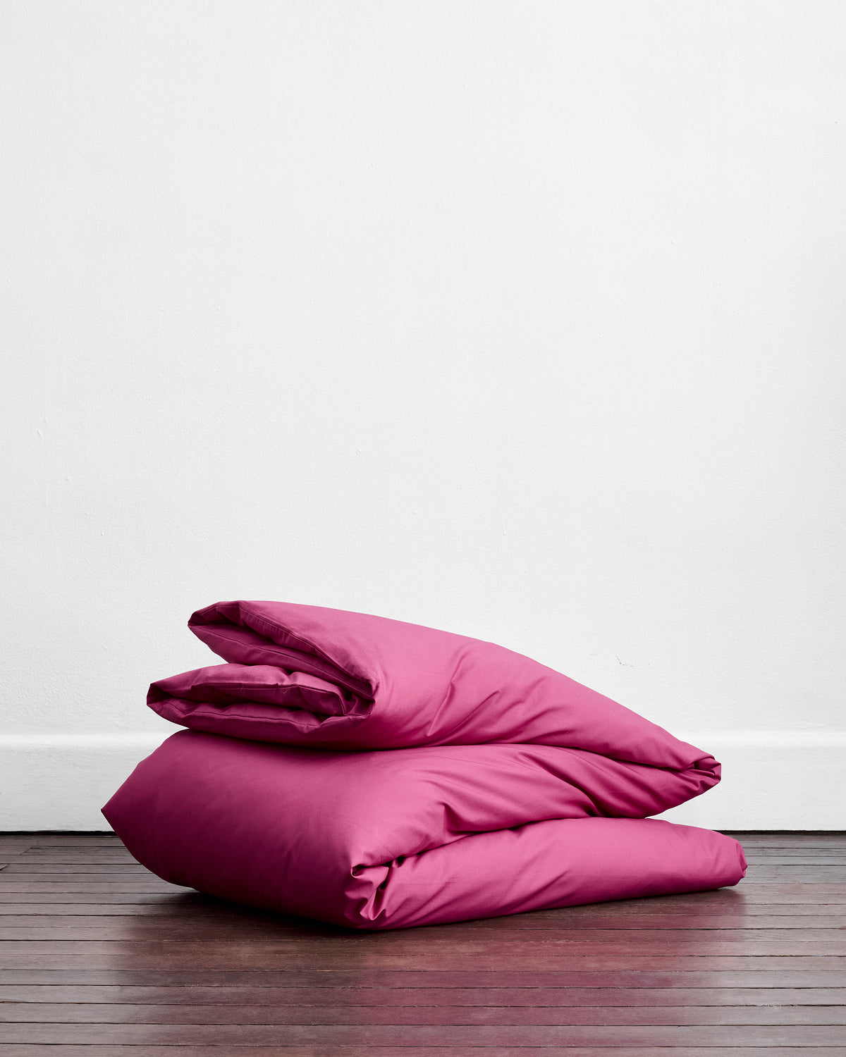 The Berry Organic Cotton Duvet Cover by Bed Threads, in bright pink, is neatly folded on a wooden floor against a plain white wall.