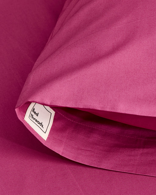 Close-up of a Berry Organic Cotton Pillowcase by Bed Threads, featuring a small white Bed Threads label. The soft, smooth fabric pillowcase is shown atop matching magenta bedding. Sold as a set of two.