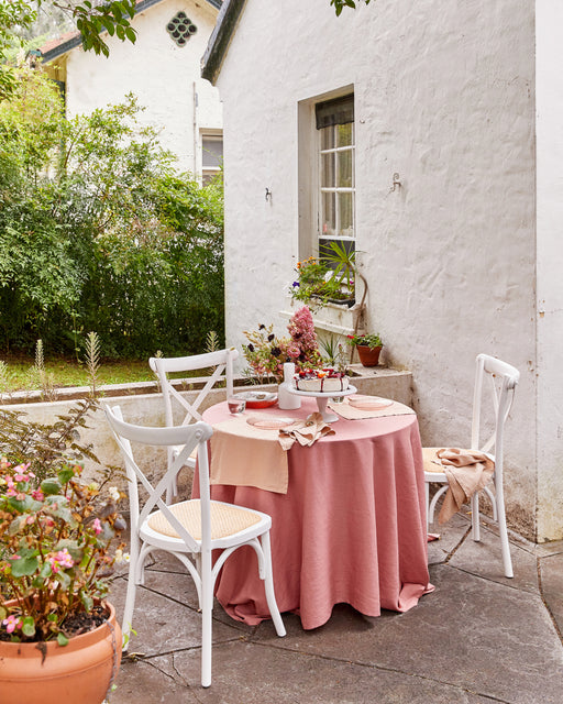 A round table with a pink tablecloth is set for three, featuring Bed Threads' Terracotta 100% French Flax Linen Placemats. White chairs circle the setup on a stone patio beside a white house, surrounded by potted plants and greenery.