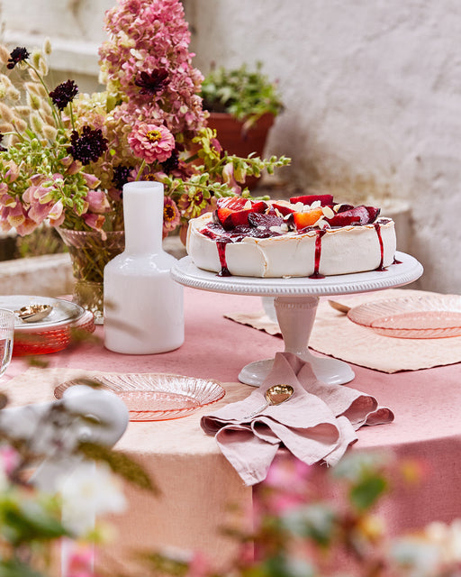 A pavlova with fresh berries sits on a white cake stand atop a pink tablecloth, surrounded by pink flowers, Bed Threads Terracotta 100% French Flax Linen Placemats (Set of Four), pink glass plates, and a white vase in an outdoor setting.