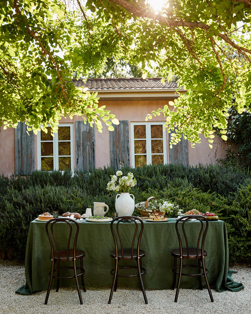 A sunlit outdoor table with a green tablecloth is set for a meal with Bed Threads' White 100% French Flax Linen Napkins (Set of Four), surrounded by four dark wood chairs under leafy branches, against a pink house with blue shutters and lush greenery.