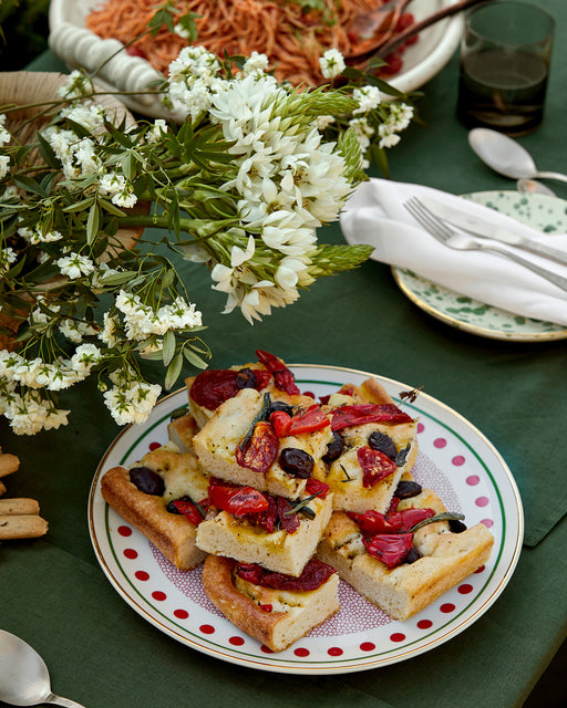 A plate of focaccia sits on the Olive 100% French Flax Linen Tablecloth by Bed Threads, beside a bowl of pasta, cutlery, a polka dot plate, and a bouquet of white flowers.