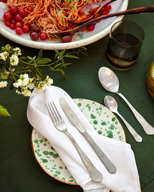 A green-spotted plate with a fork, knife, and Bed Threads White 100% French Flax Linen Napkins (Set of Four) sits on a green tablecloth beside a glass, spoon, and a large serving bowl of tomato spaghetti. White flowers decorate the table.