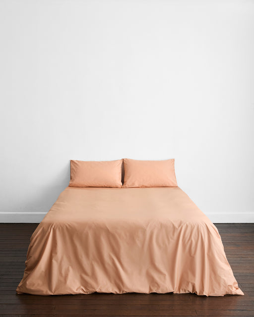 A neatly made bed features Bed Threads’ Butterscotch Organic Cotton Pillowcases (Set of Two) in a peach hue, paired with matching sheets. Centered against a plain white wall on a dark wooden floor, the minimalistic room is free of decorations or extra furniture.