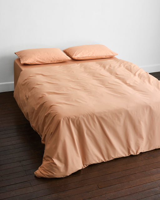 A neatly made bed features the Butterscotch Organic Cotton Bedding Set by Bed Threads, including two matching pillows, set against a white wall on a dark wooden floor.