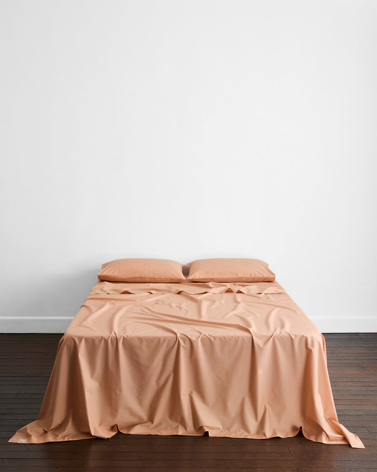 A neatly made bed with peach-colored sheets and two Bed Threads Butterscotch Organic Cotton King Pillowcases rests against a plain white wall on a dark wooden floor.