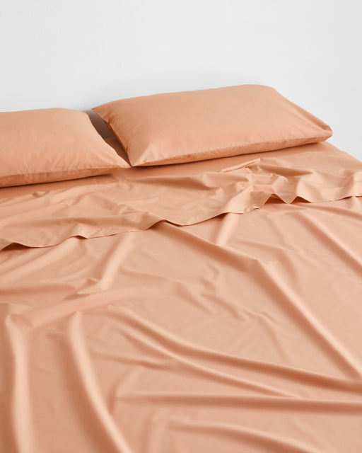 A neatly made bed with two pillows and a wrinkled Butterscotch Organic Cotton Fitted Sheet by Bed Threads, set against a plain white wall.