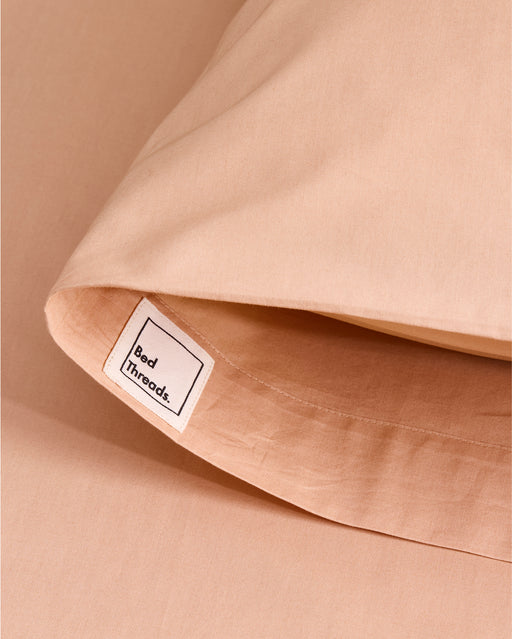 A close-up of the Butterscotch Organic Cotton Pillowcases (Set of Two) by Bed Threads shows smooth, peach-toned percale fabric with a white Bed Threads label, neatly stitched and resting on a matching sheet.