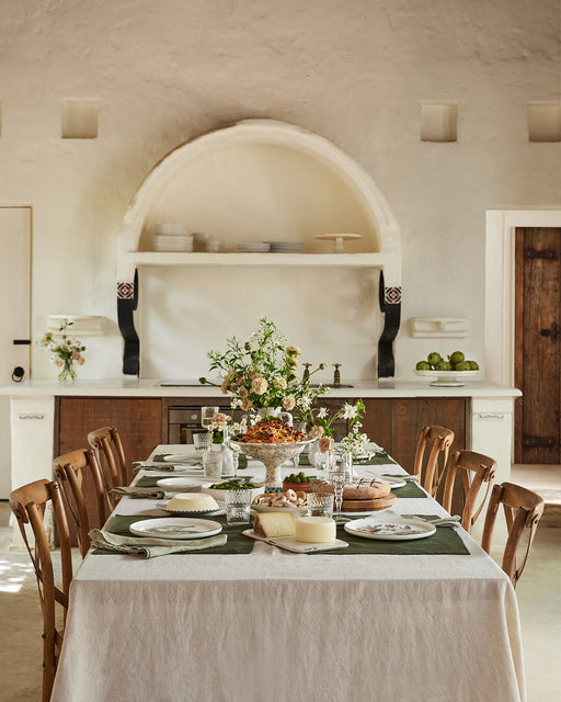 A rustic kitchen features a long dining table set with a white tablecloth, Bed Threads Olive 100% French Flax Linen Placemats (Set of Four), a green runner, floral centerpiece, plates, glasses, and bread. Arched shelf and wooden cabinets are visible behind.