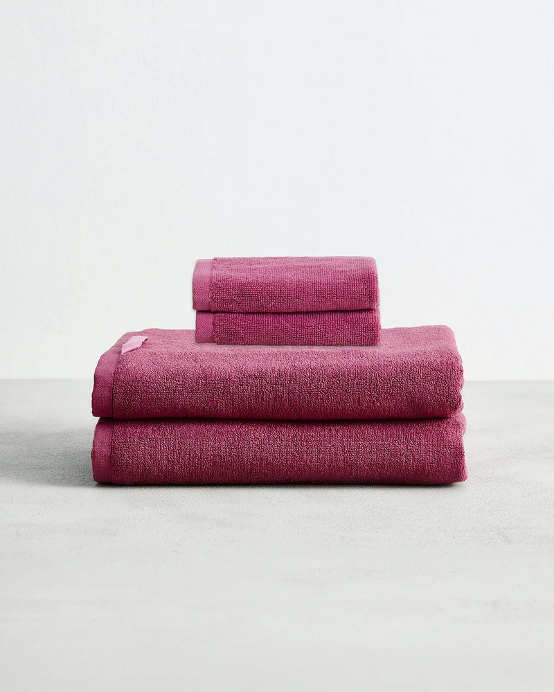 Set of two berry pink face washers and two hand towels in 100% cotton terry stacked on a light grey surface with a white background. 