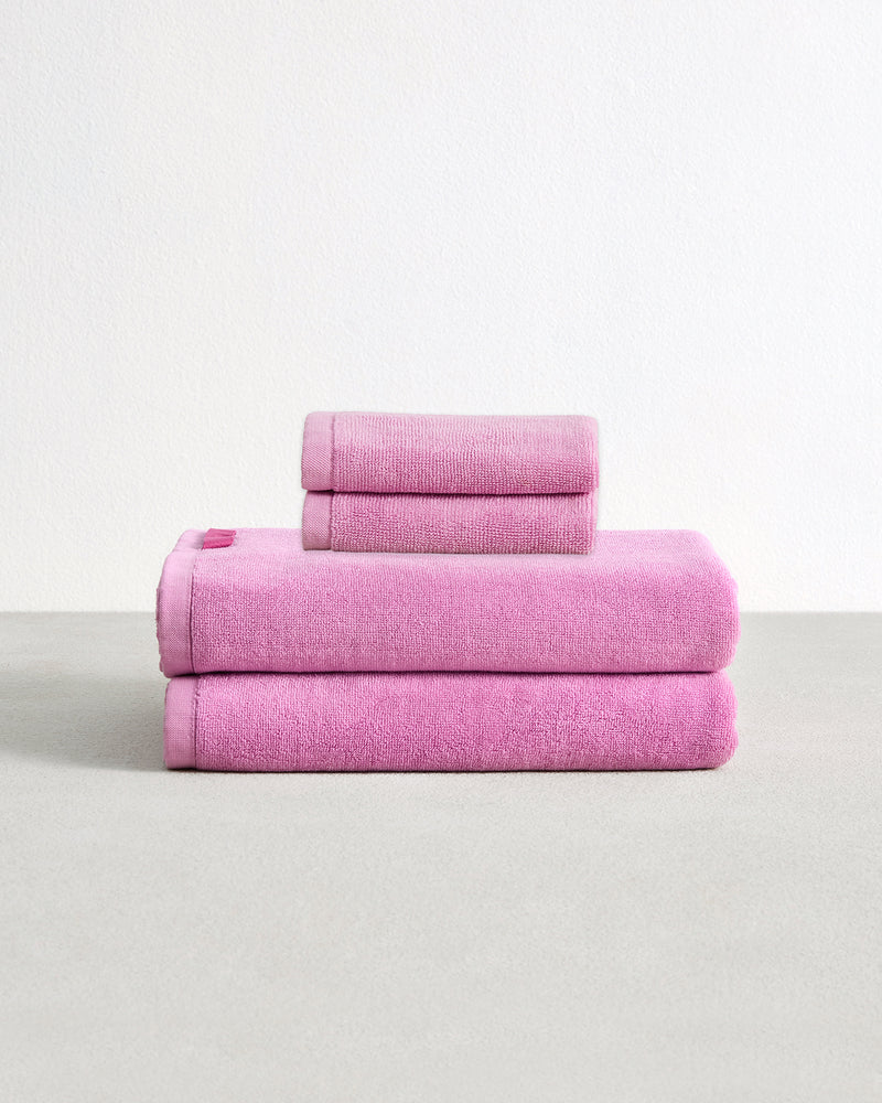 Set of two bubblegum pink face washers and two hand towels in 100% cotton terry stacked on a light grey surface with a white background. 