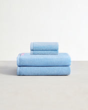 Set of two sky blue face washers and two bath towels in 100% cotton terry stacked on a light grey surface with a white background.  - view