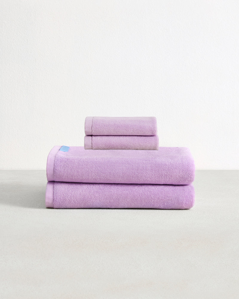 Set of two violet purple face washers and two bath towels in 100% cotton terry stacked on a light grey surface with a white background. 