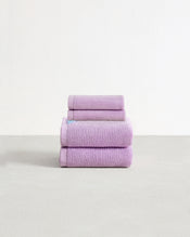 Set of two violet purple face washers and two hand towels in 100% cotton terry stacked on a light grey surface with a white background.  - view