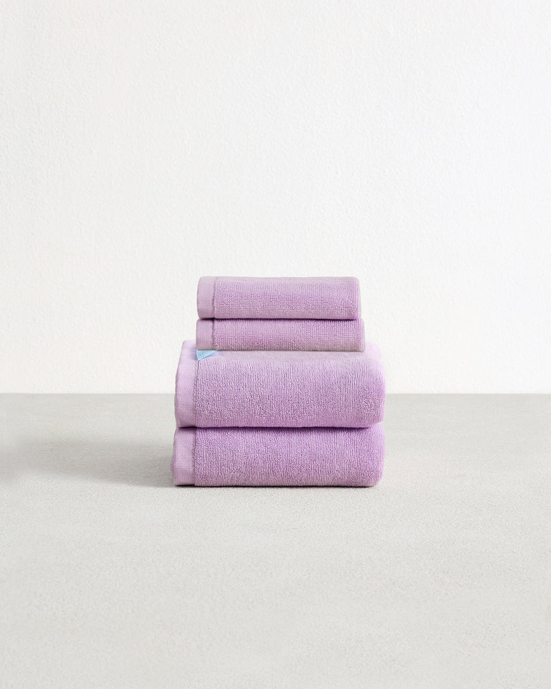 Set of two violet purple face washers and two hand towels in 100% cotton terry stacked on a light grey surface with a white background. 