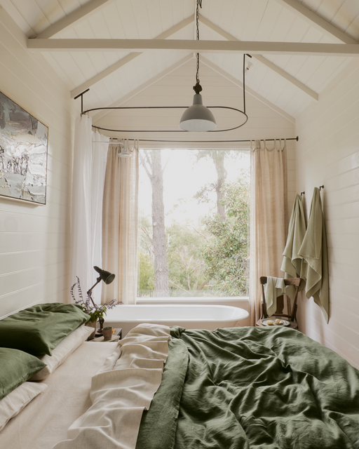A cozy bedroom features Bed Threads' Olive & Oatmeal & White Stripe 100% French Flax Linen Bedding Bundle, a lamp on the nightstand, beige curtains framing a large window with greenery outside, towels on the wall, and a chair by the tub.