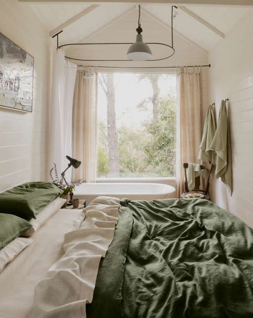 A cozy bedroom features Bed Threads’ Oatmeal & White Stripe 100% French Flax Linen King Pillowcases, green and beige bedding, sheer curtains framing a large window, a bathtub below, soft light, potted plant, wall towels, and lush greenery outside.