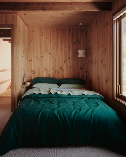 A cozy bedroom with wood-paneled walls, a bed featuring the Forest 100% French Flax Linen Duvet Cover by Bed Threads and green pillows, a small floating nightstand with water bottles, a white pendant lamp, and a window letting in natural light.
