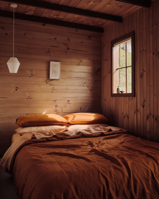 A cozy bedroom with wood-paneled walls features a bed dressed in orange bedding and Bed Threads' Rust 100% French Flax Linen Pillowcases, a small window with natural light, a white pendant lamp, and a framed picture above the bed.