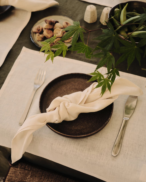 A brown plate with a knotted cream napkin, fork, and knife sits atop Bed Threads' Oatmeal 100% French Flax Linen Placemats (Set of Four). Green leaves, figs, and salt and pepper shakers accent the elegant table setting.