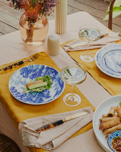 A table set with Bed Threads Turmeric 100% French Flax Linen Placemats, blue and white plates, gold cutlery, wine glasses, flowers, candles, and spring rolls creates a bright, elegant scene that brings style and protection to your meals.