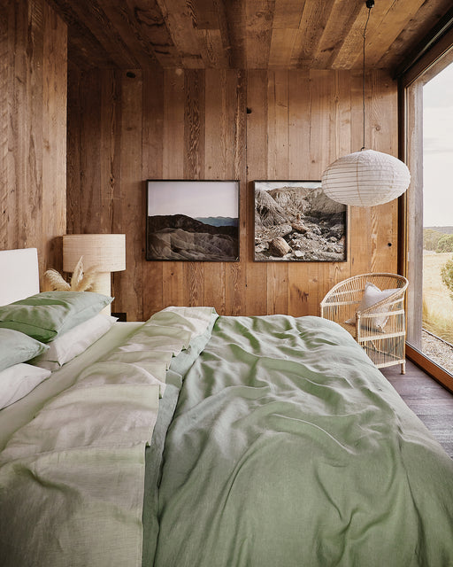 A cozy bedroom features wood-paneled walls, a Bed Threads Sage 100% French Flax Linen Fitted Sheet, a wicker chair, paper lantern, and large windows with scenic views. Two framed landscapes hang above a small table and lamp.