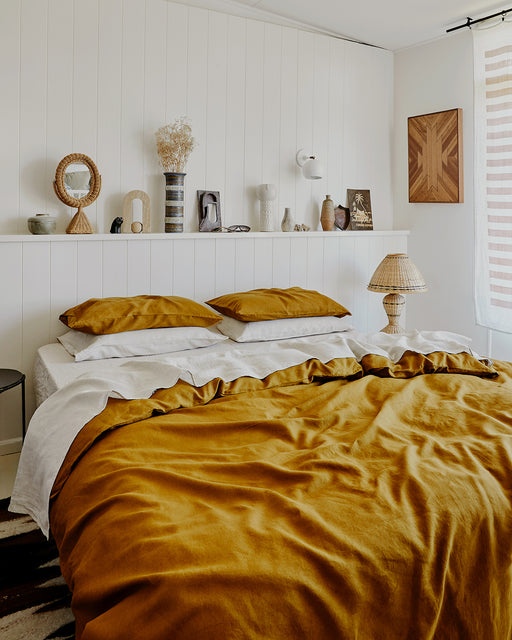 A cozy bedroom featuring the Bed Threads Turmeric 100% French Flax Linen Duvet Cover Set with matching pillowcases, white walls, decorative vases on a shelf, art, and a rattan lamp by the bed. Sheer curtains let in natural light.