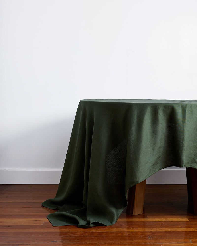 A table featuring the Bed Threads Olive 100% French Flax Linen Tablecloth, its rich green fabric partially concealing the wooden legs, stands on a polished wood floor against a plain white wall.