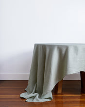 The Bed Threads Sage 100% French Flax Linen Tablecloth in light gray drapes over a round table, pooling onto wood flooring. A plain white wall and baseboard complete this elegant linen table setting. - view