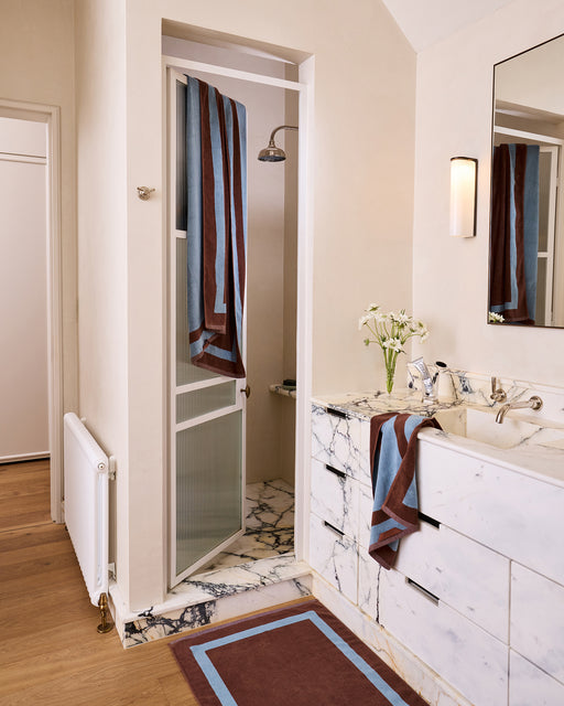 A woman sits on the edge of a white bathtub, wrapped in a Bed Threads Cacao & Lagoon Two-Toned 100% Cotton Terry Bath Towel, with a matching towel on her head. A rug, stool with toiletries, and fresh flowers complete the scene.