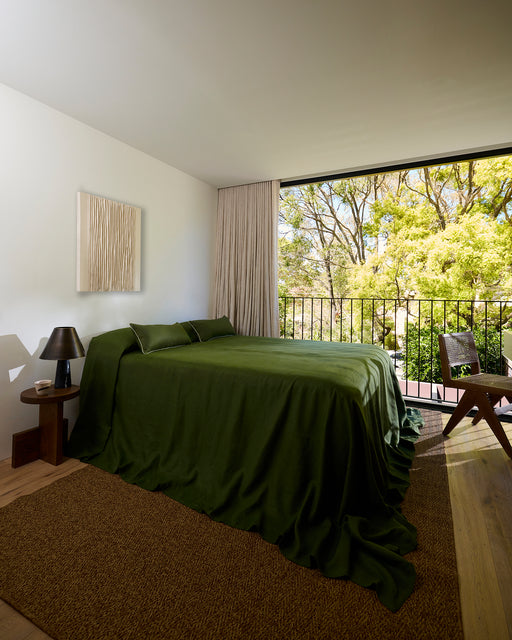 A neatly made bed with sage green bedding, two light green pillows with dark green piping, and a Bed Threads Olive & Sage 100% French Flax Linen Piped Lumbar Cushion Cover in dark green in front.