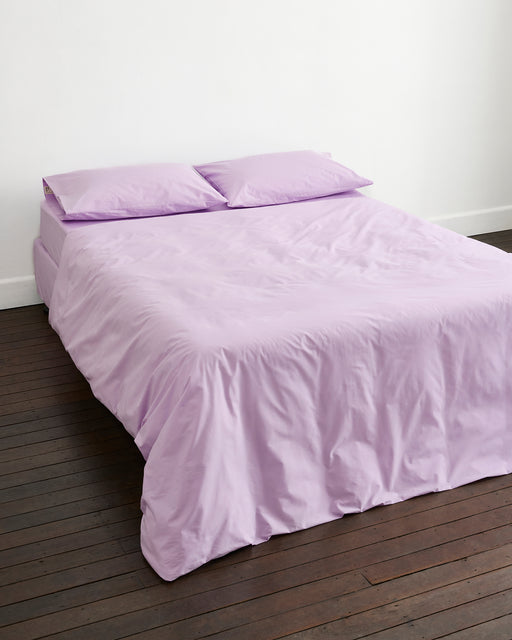 A neatly made bed with lavender sheets and a Bed Threads Violet 100% Organic Cotton Duvet Cover sits against a white wall on a dark wooden floor, accented by two matching pillows.