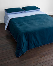 A neatly made bed features the Bed Threads Aegean, Sky & Bubblegum Piped 100% Organic Cotton Duvet Cover with matching dark blue percale bedding and pillows, all set against a white wall on a dark wooden floor. - view