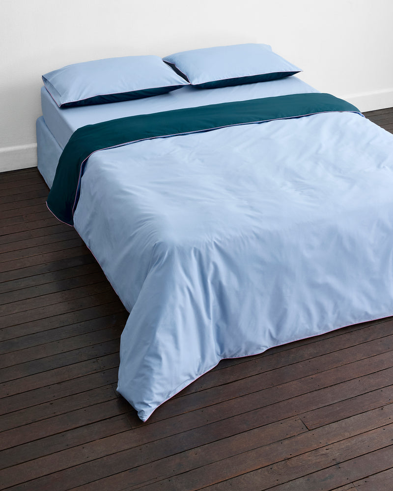 A neatly made bed features Bed Threads’ Aegean, Sky & Bubblegum Piped 100% Organic Cotton Duvet Cover atop light blue percale sheets and pillowcases, with a dark teal blanket on a dark wooden floor against a plain white wall.
