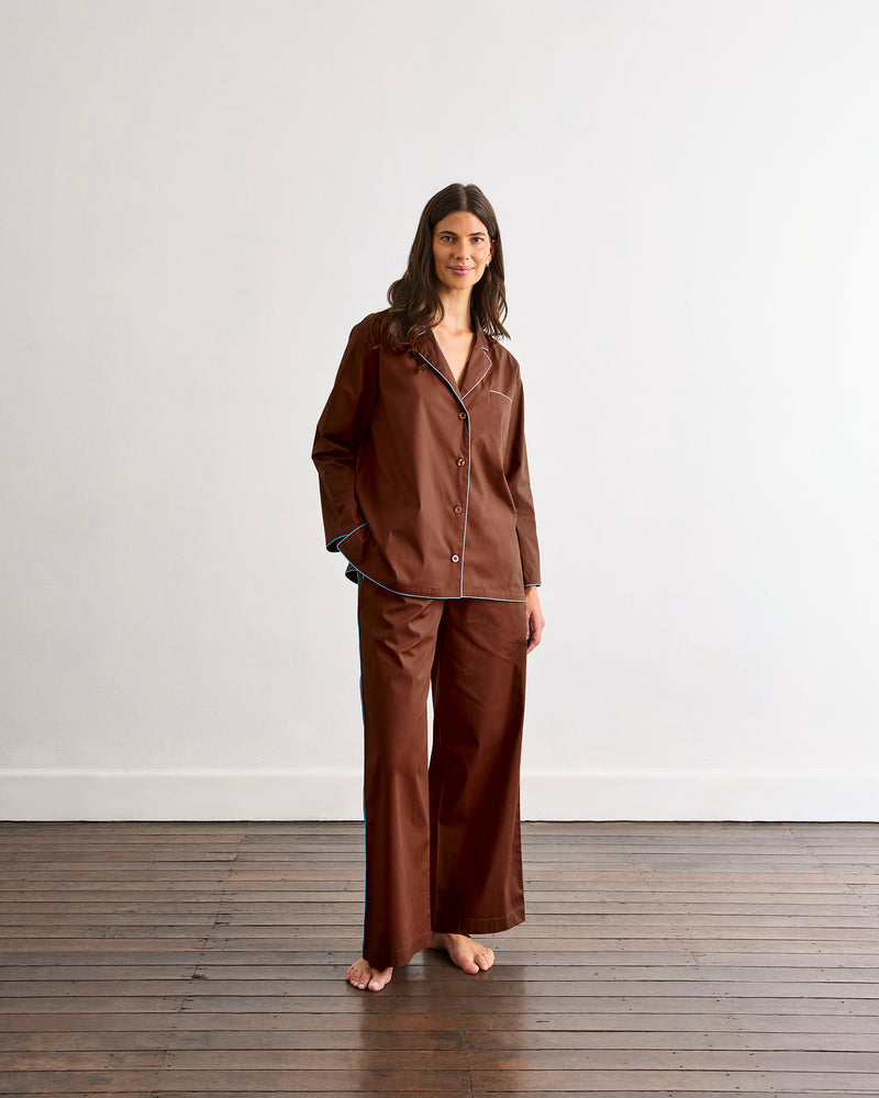 A woman stands barefoot on a wooden floor in the Bed Threads Espresso & Sky Piped Long Sleeve Shirt, a brown, loose-fitting cotton top with subtle contrast piping, set against a plain white wall.
