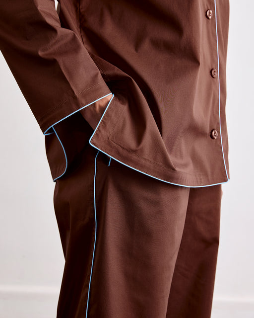 A person wearing Bed Threads' Espresso & Sky Piped Pants, shown from shoulders to knees, features brown relaxed-fit pants with light blue piping and an elasticated waist, one hand in their pocket.