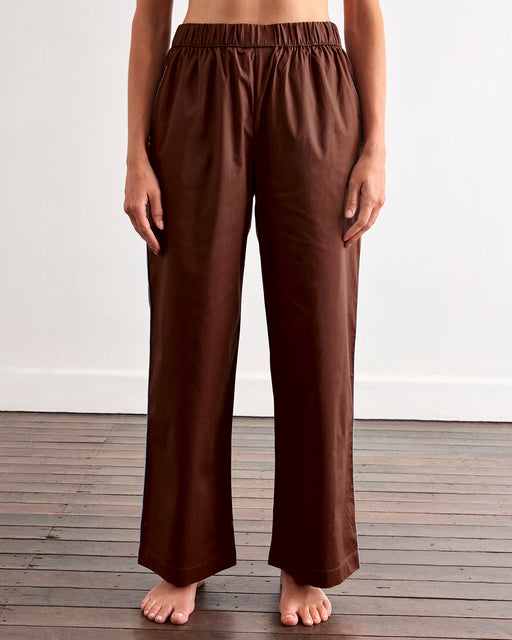 A woman with long brown hair stands barefoot on a wooden floor against a white wall, wearing Bed Threads' Espresso & Sky Piped Pants—a brown pajama set with white piping and relaxed, elastic-waist pants.