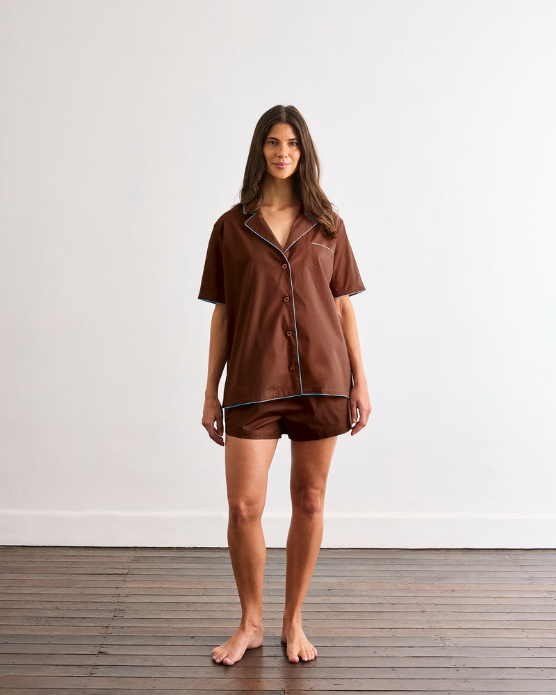 A person wearing the Bed Threads Espresso & Sky Piped Short Sleep Set, featuring brown piped shorts with an elastic waistband and a short sleeve shirt, shown from waist to mid-thigh against a neutral background.