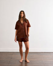 A woman stands barefoot on a wooden floor against a plain white wall, wearing Bed Threads’ Espresso & Sky Piped Shorts pajama set with white piping and breathable cotton fabric. - view