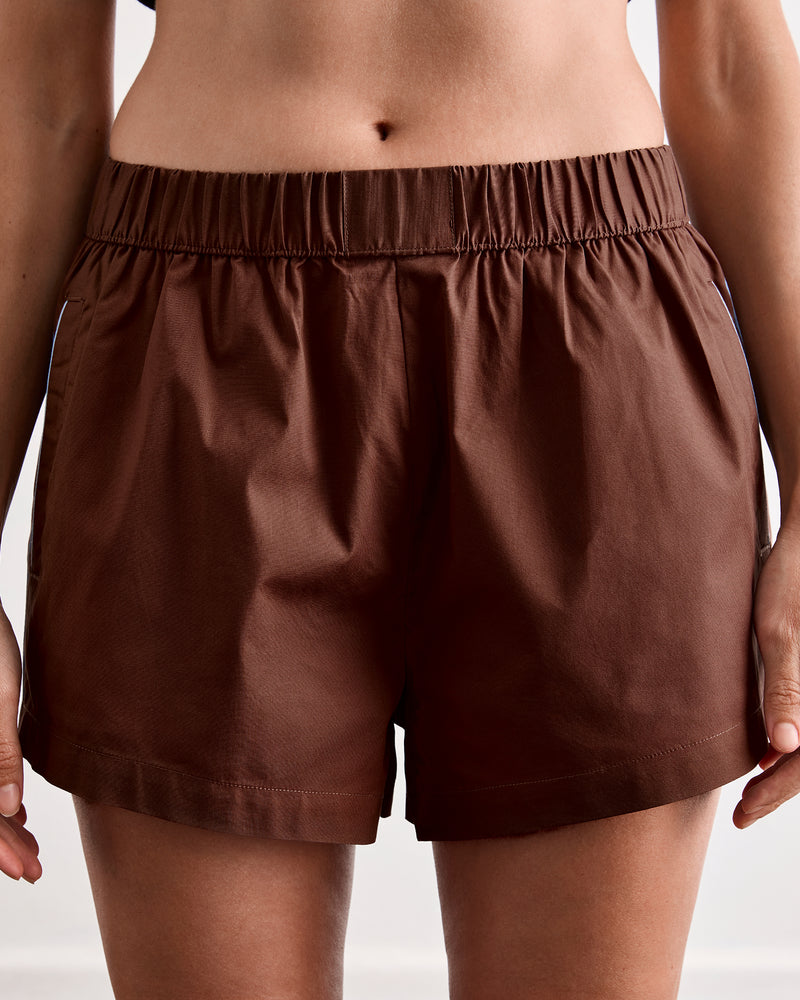 A woman stands barefoot on a wooden floor against a plain white wall, wearing Bed Threads’ Espresso & Sky Piped Shorts pajama set with white piping and breathable cotton fabric.