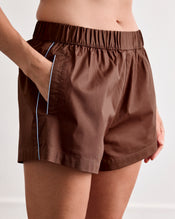 A person wears Bed Threads' Espresso & Sky Piped Shorts, featuring an elastic waistband and breathable cotton with a thin white stripe along the pocket edge. - view