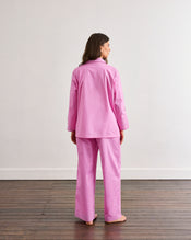 A woman stands barefoot on wooden floorboards, facing away from the camera, wearing the Bed Threads Bubblegum & Berry Piped Long Sleeve Shirt in a plain room with white walls. - view